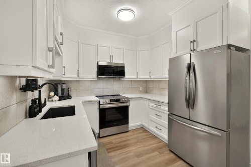 Modern kitchen featuring white cabinetry, light-toned countertops, stainless steel appliances, a tiled backsplash, and wood-finish flooring - #9 175 Mcconachie Dr Nw, Edmonton, AB 
