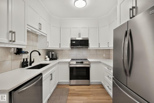 U-shaped kitchen featuring white cabinetry, light-toned countertops, tile backsplash, stainless steel appliances, and wood-finish flooring - #9 175 Mcconachie Dr Nw, Edmonton, AB 