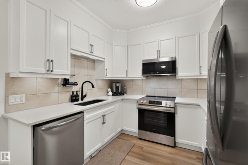 White cabinetry, stainless steel appliances, light-toned tiled backsplash, solid surface countertops, and wood-finish flooring - #9 175 Mcconachie Dr Nw, Edmonton, AB 
