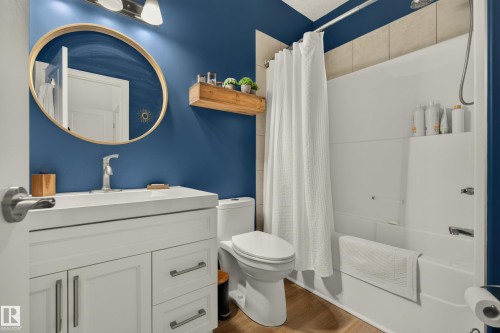 White vanity with brushed nickel hardware, circular mirror with gold-tone frame, white bathtub with shower, light wood-finish flooring, and a floating wood shelf - #9 175 Mcconachie Dr Nw, Edmonton, AB 