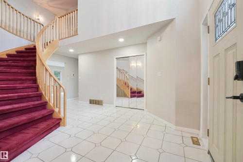 Grand entryway featuring a curved staircase with wood banister and red carpeted treads, white tile flooring with geometric accents, recessed lighting, and a front door with decorative glass inlay - 5107 17 Avenue Nw, Edmonton, AB 
