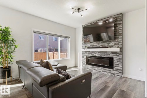 Living area featuring wood-finish flooring, a contemporary ceiling light fixture, a large window, and a fireplace with a stone-clad surround and a floating white mantel - 19703 29 Avenue Nw, Edmonton, AB 