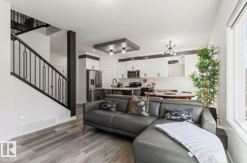 Open-concept living area featuring wood-finish flooring, a modern staircase with black railings, and white kitchen cabinetry with stainless steel appliances - 19703 29 Avenue Nw, Edmonton, AB 