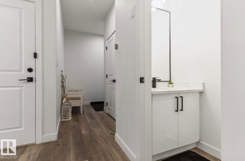Wood-finish flooring, white paneled doors with matte black hardware, white vanity with a rectangular sink, matte black faucet, and a wall-mounted mirror - 19703 29 Avenue Nw, Edmonton, AB 