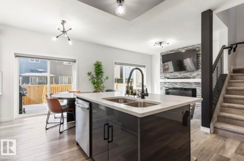 Kitchen island with integrated sink, wood-finish flooring, stone-clad fireplace, black metal stair railing, and sliding glass doors to outdoor space - 19703 29 Avenue Nw, Edmonton, AB 