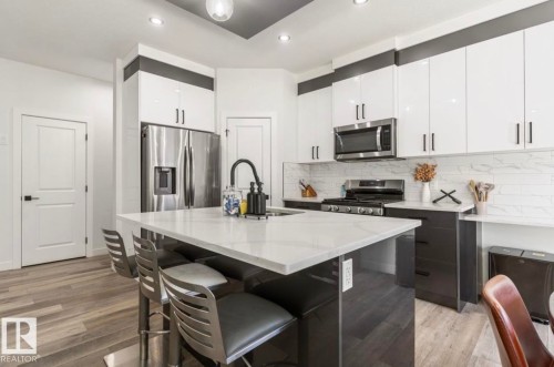 Kitchen with wood-finish flooring, a central island with a light-colored countertop, white upper cabinetry, dark lower cabinetry, and stainless steel appliances - 19703 29 Avenue Nw, Edmonton, AB 