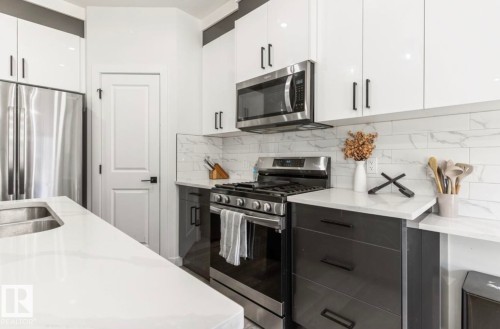 Modern kitchen featuring stainless steel appliances, white upper cabinetry, dark lower cabinetry, white stone countertops, and a white tile backsplash - 19703 29 Avenue Nw, Edmonton, AB 