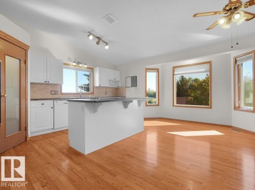 Spacious room featuring wood-finish flooring, white cabinetry, a kitchen island with a dark countertop, multiple windows with wood trim, and a ceiling fan with integrated lighting - 535 Pardee Bay Nw, Edmonton, AB 
