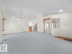 Expansive carpeted area with vaulted ceilings, a central chandelier, wood-trimmed windows, and a staircase - 