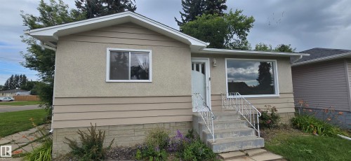 Property exterior featuring light-toned stucco and horizontal siding, a prominent front entry with concrete steps and a metal handrail, and white-framed windows - 4818 54A Avenue, Vegreville, AB 