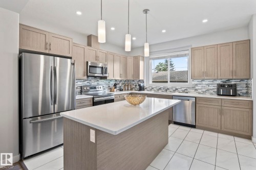 Contemporary kitchen featuring wood-finish cabinetry, stainless steel appliances, a center island with a light-toned countertop, and a tiled backsplash - 10338 162St Nw, Edmonton, AB 