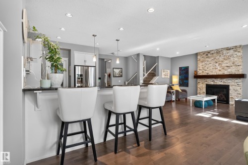Open-concept living space featuring wood-finish flooring, a stone-clad fireplace with a wood mantle, recessed lighting, a breakfast bar, and a visible staircase - 22933 94A Avenue Nw, Edmonton, AB 