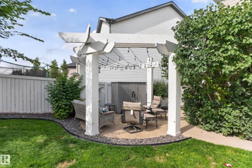 Outdoor living area featuring a white pergola, gravel patio, string lighting, and surrounding green landscaping - 22933 94A Avenue Nw, Edmonton, AB 