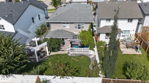 Expansive backyard featuring a manicured lawn, established landscaping, white pergola-covered patio, paved walkway, and a rear deck with white railings - 22933 94A Avenue Nw, Edmonton, AB 