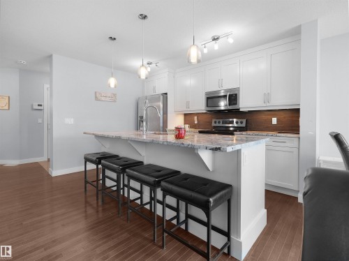 Kitchen featuring white shaker cabinetry, stainless steel appliances, a wood-finish backsplash, a kitchen island with a stone-finish countertop, and wood-finish flooring - 6656 Cardinal Rd Sw, Edmonton, AB 
