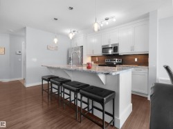Kitchen featuring white shaker cabinetry, stainless steel appliances, a wood-finish backsplash, a kitchen island with a stone-finish countertop, and wood-finish flooring - 