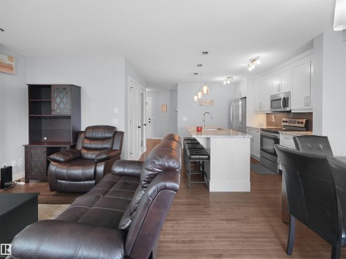 Open-concept living space with wood-finish flooring, white cabinetry, stainless steel appliances, and a kitchen island with pendant lighting - 6656 Cardinal Rd Sw, Edmonton, AB 