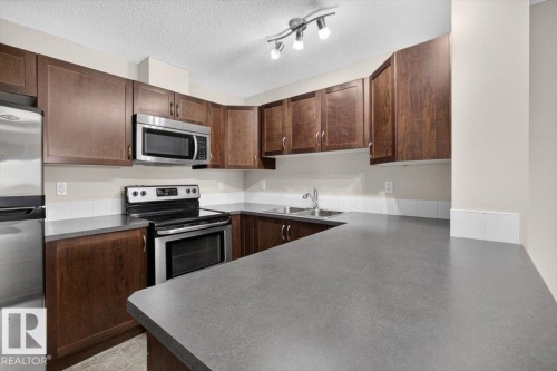 Kitchen featuring dark wood-finish cabinetry, stainless steel appliances, a double basin sink, track lighting, and grey countertops with a white tile backsplash - #316 392 Silver Berry Rd Nw, Edmonton, AB - Indoor Photo Showing Kitchen With Double Sink