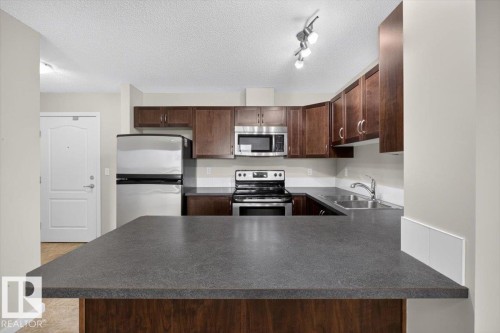 Kitchen featuring dark wood-finish cabinetry, stainless steel appliances, dark countertops, and a track lighting fixture - #316 392 Silver Berry Rd Nw, Edmonton, AB - Indoor Photo Showing Kitchen With Double Sink