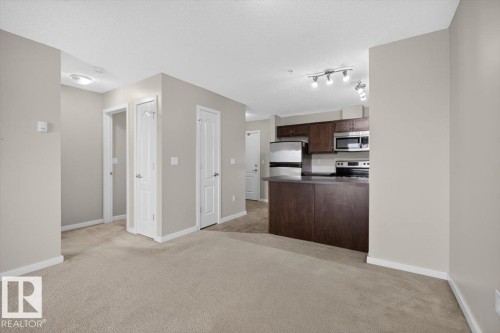 Open concept living area with carpeting, featuring a kitchen peninsula with a dark countertop, stainless steel appliances, dark wood-finish cabinetry, and track lighting - #316 392 Silver Berry Rd Nw, Edmonton, AB - Indoor Photo Showing Kitchen