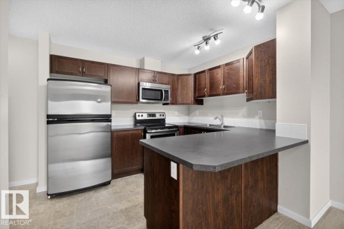 Kitchen featuring stainless steel appliances, dark wood-finish cabinetry, and a multi-level countertop island - #316 392 Silver Berry Rd Nw, Edmonton, AB - Indoor Photo Showing Kitchen With Double Sink