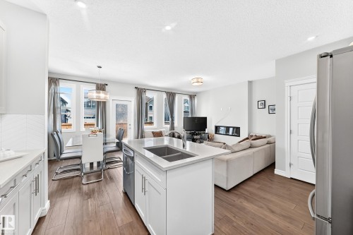 Open-concept living space featuring wood-finish flooring, a kitchen island with an integrated sink, white cabinetry with silver-tone hardware, and a linear fireplace - 2628 Donaghey Crescent Sw, Edmonton, AB 