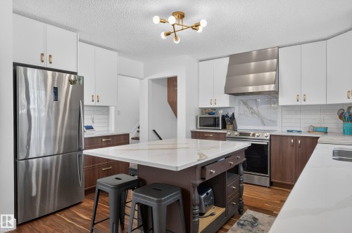 Modern kitchen featuring white upper cabinetry, wood-finish lower cabinetry, a center island with a light-toned countertop, stainless steel appliances, and wood-finish flooring - 18316 71 Avenue Nw, Edmonton, AB 