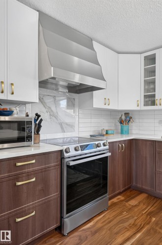 Modern kitchen featuring a stainless steel range with a flat cooktop, custom range hood, white upper cabinetry, rich wood-finish lower cabinetry, and a white subway tile backsplash - 18316 71 Avenue Nw, Edmonton, AB 