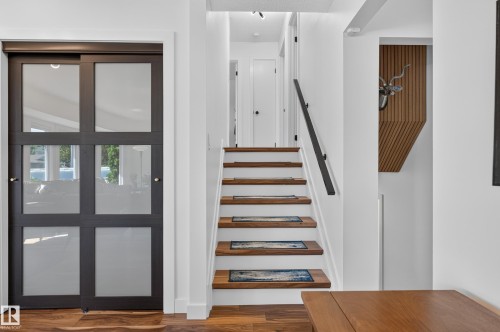 Dark-framed sliding glass door, rich wood-finish flooring, staircase with wood-finish treads and risers, black metal handrail, and a recessed wood-slat accent wall - 18316 71 Avenue Nw, Edmonton, AB 