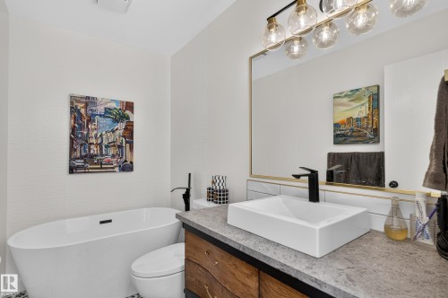 Freestanding soaking tub, wood-finish vanity with a rectangular vessel sink, matte black faucet, and a large framed mirror with globe lighting - 18316 71 Avenue Nw, Edmonton, AB 