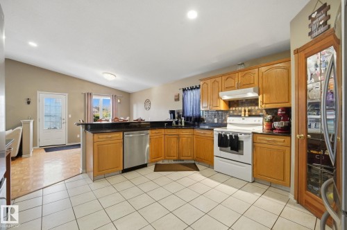 Kitchen featuring light wood cabinetry, black countertops, white tile flooring, stainless steel dishwasher, and white range - 5132 157 Avenue Nw, Edmonton, AB 
