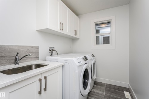 Laundry area featuring upper cabinetry, a utility sink with a stone backsplash, a window, and tiled flooring - 9107 181 Avenue Nw, Edmonton, AB 