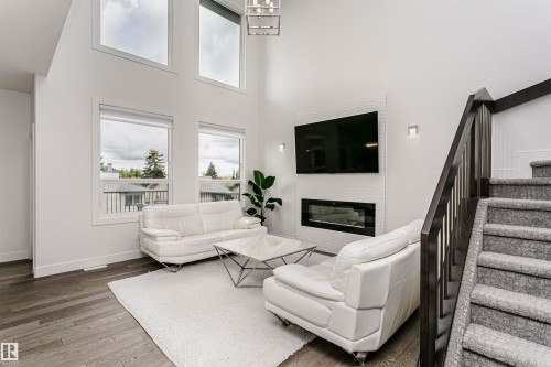Living area featuring a modern linear fireplace with a textured tile surround, high ceilings, large windows, wood-finish flooring, and a dark wood staircase with carpeted treads - 9107 181 Avenue Nw, Edmonton, AB 