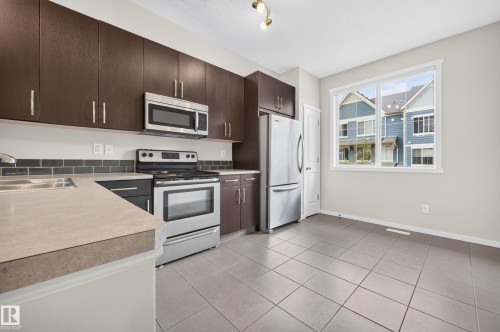 Kitchen featuring dark wood-finish cabinetry, stainless steel appliances, light-toned countertops, a subway tile backsplash, and ceramic tile flooring - #72 603 Watt Bv Sw Sw, Edmonton, AB 