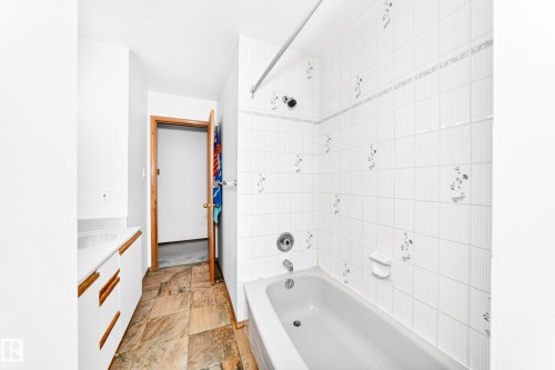 Built-in bathtub with white tile surround, white vanity cabinetry with wood-finish handles, and stone-finish flooring - 4208 Ramsay Road Nw, Edmonton, AB 