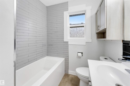 White bathtub with gray horizontal tile surround, wood-finish wall-mounted cabinetry, white vanity with integrated sink, and a framed window - 7928 82 Avenue Nw, Edmonton, AB 