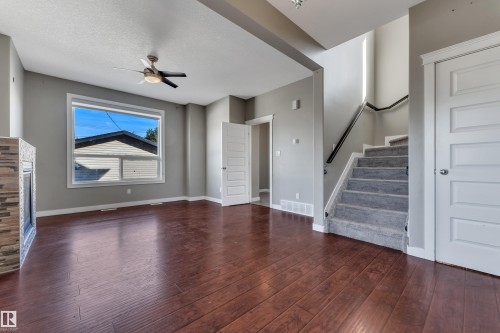 Wood-finish flooring, a ceiling fan with integrated lighting, a stone-clad fireplace, and a carpeted staircase with a dark handrail - 11421 82 Street Nw, Edmonton, AB 