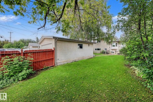 Expansive green lawn with mature trees and shrubs, featuring a detached garage with stucco exterior, and a red wood fence - 11431 51 Avenue Nw, Edmonton, AB 