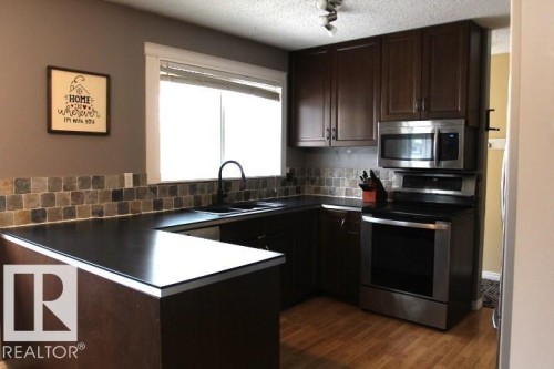 Kitchen featuring dark wood-finish cabinetry, dark countertops, a tiled backsplash, and stainless steel appliances - 64 Apache Crescent, Leduc, AB 
