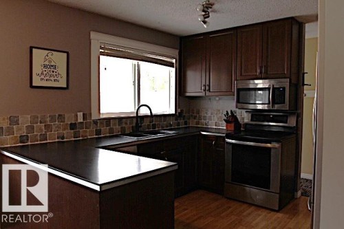 U-shaped countertop with dark finish, wood-finish flooring, dark wood cabinetry, stainless steel microwave, and stainless steel range - 64 Apache Crescent, Leduc, AB 