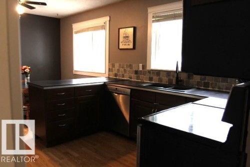 Wood-finish flooring, dark cabinetry with bar pull hardware, L-shaped countertop, tile backsplash, and a ceiling fan with light fixture - 64 Apache Crescent, Leduc, AB 