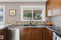 Kitchen featuring wood cabinetry, dark countertops, square tile backsplash, and a stainless steel dishwasher - 