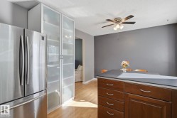 Stainless steel refrigerator, integrated pantry cabinetry with frosted glass panels, wood-finish flooring, ceiling fan with integrated lighting, and a kitchen island with dark wood-finish cabinetry and a contrasting countertop - 