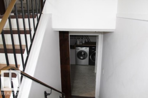 Stairwell with wood treads and black metal balusters, featuring a laundry area with a washer and dryer visible through a doorway - 64 Apache Crescent, Leduc, AB 