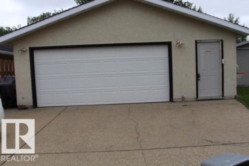 Attached two-car garage with a white panel door and a side entry door, featuring a stucco exterior and concrete driveway - 64 Apache Crescent, Leduc, AB 