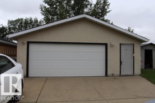 Stucco exterior featuring a white paneled garage door, a side entry door, exterior lighting fixtures, and a paved driveway - 64 Apache Crescent, Leduc, AB 