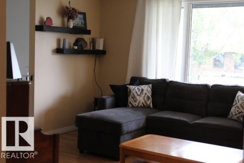 Bright living room showcasing a black sectional sofa with geometric patterned accent pillows, wood-finish flooring, and abundant natural light from large windows - 64 Apache Crescent, Leduc, AB 