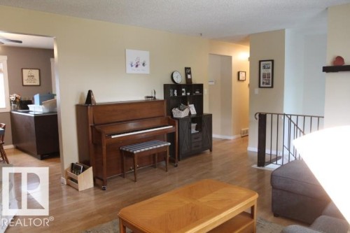 Open-concept living area featuring warm wood-finish flooring throughout, a wood upright piano with bench, black cabinetry with shelving, wood coffee table, and abundant natural light from multiple windows - 64 Apache Crescent, Leduc, AB 
