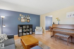 Spacious living area showcasing warm wood-finish flooring, blue accent wall with decorative shelving, black built-in cabinetry with open shelving, cream-colored upholstered seating, and a wood upright piano - 