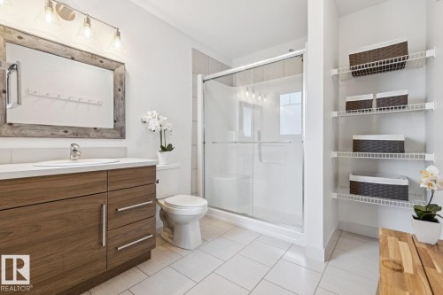 White tile flooring, wood-finish vanity with countertop, framed mirror, glass-enclosed shower, and wall-mounted shelving - 22434 99A Avenue Nw, Edmonton, AB 